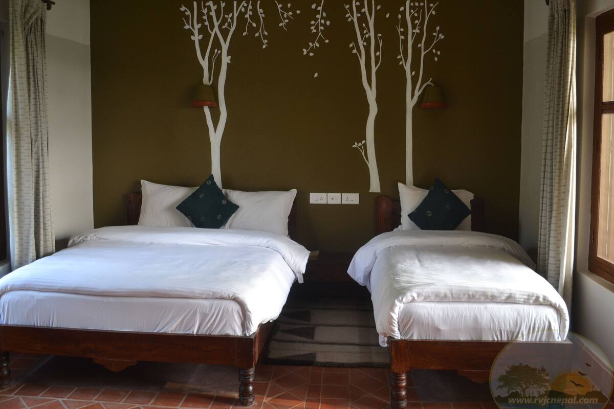 A room with two beds, each with white bedding and dark pillows. There are wall decorations of white trees and a small shelf with items.