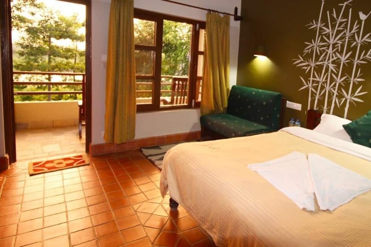 A hotel room with a bed, a green chair, a balcony with a view, and a wall-mounted lamp.