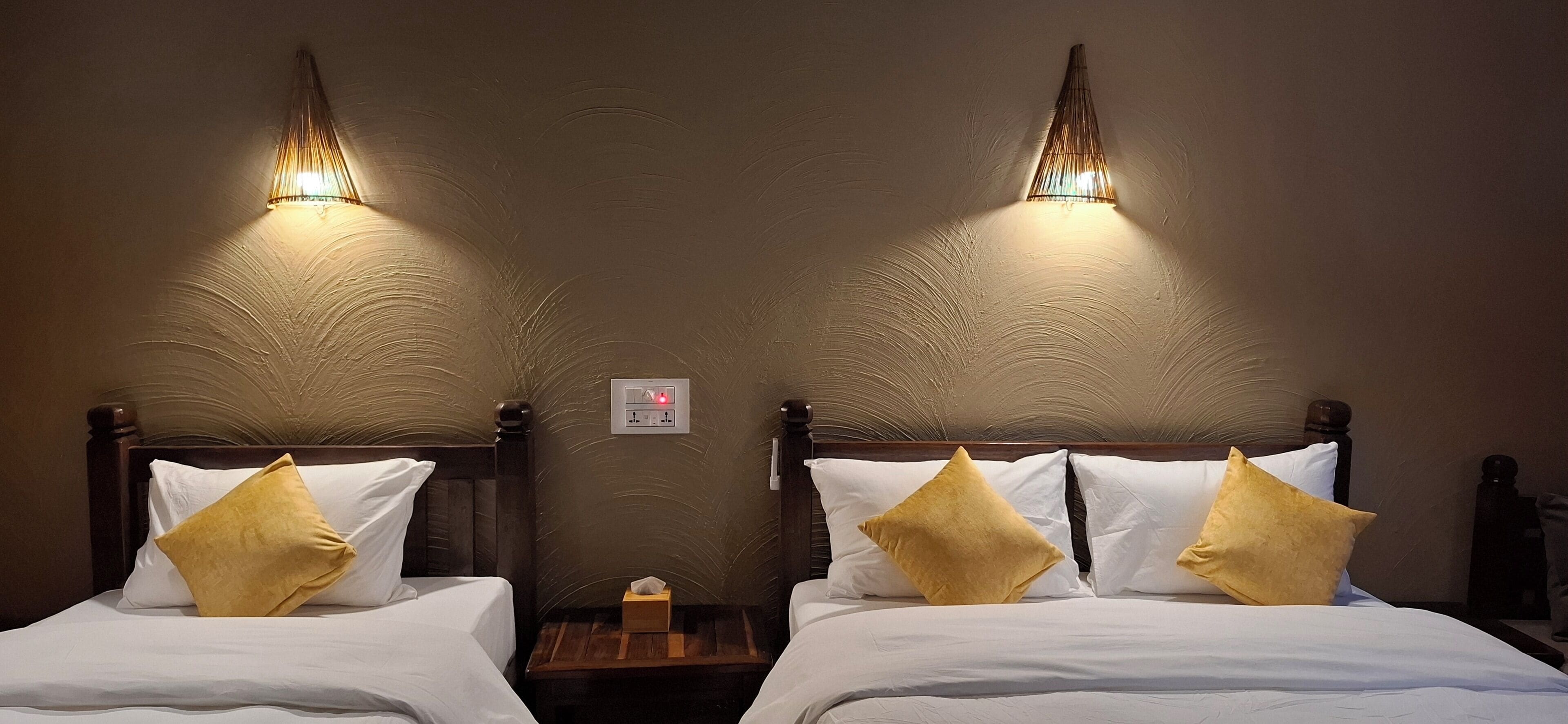 Dos camas dobles con sábanas blancas y almohadas doradas, cada una con cabecera de madera y una mesita de noche.