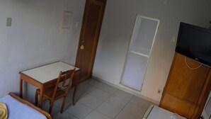Una habitación pequeña y poco amoblada, con una mesa y una silla de madera, una puerta cerrada, un ventanal con persiana y un televisor sobre un mueble de madera.