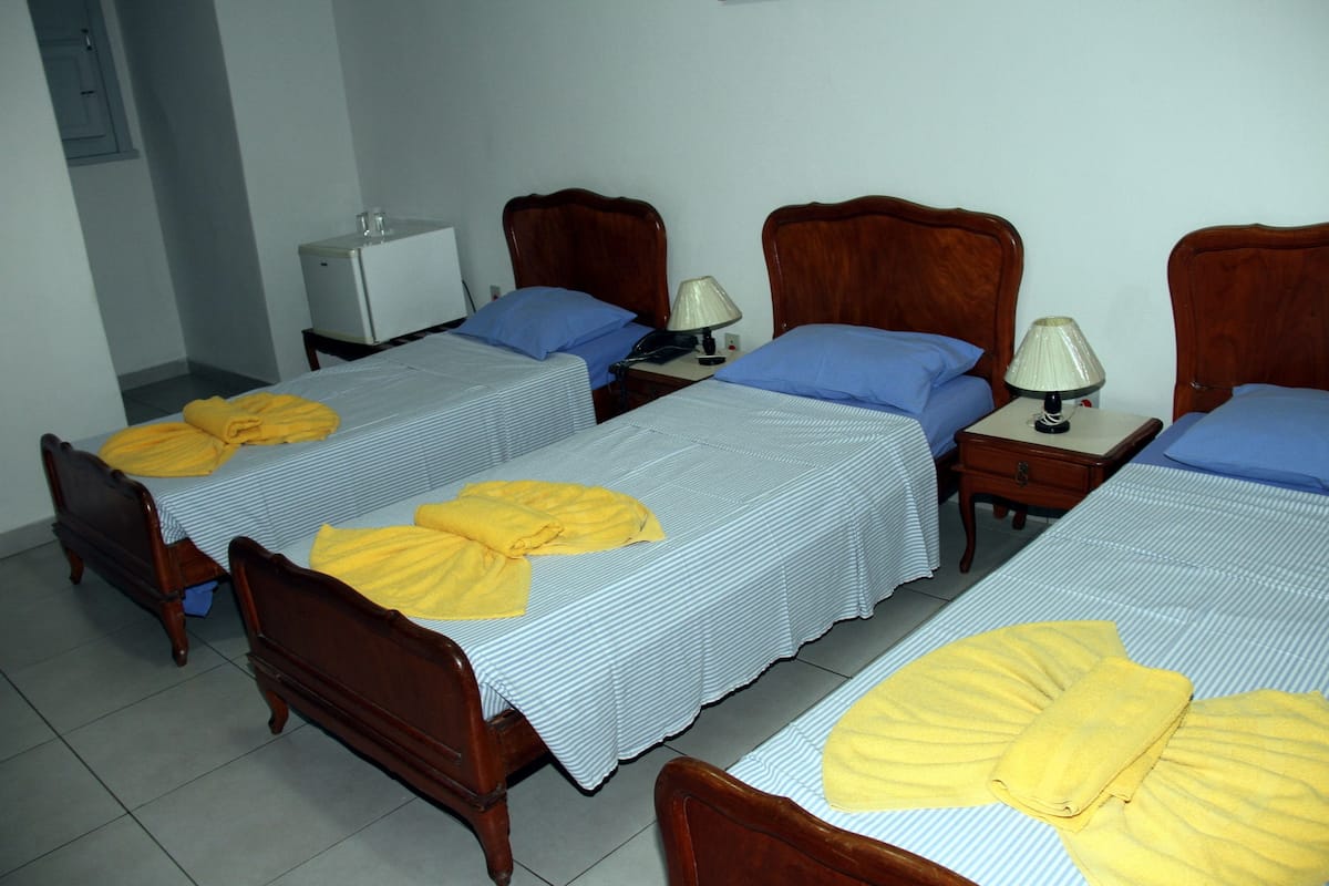 Une chambre d’hôtel avec quatre lits simples, chacun doté d’une tête de lit en bois et d’une couvre-lit à rayures. On y trouve deux tables de chevet avec des lampes, ainsi qu’un petit réfrigérateur blanc dans un coin.