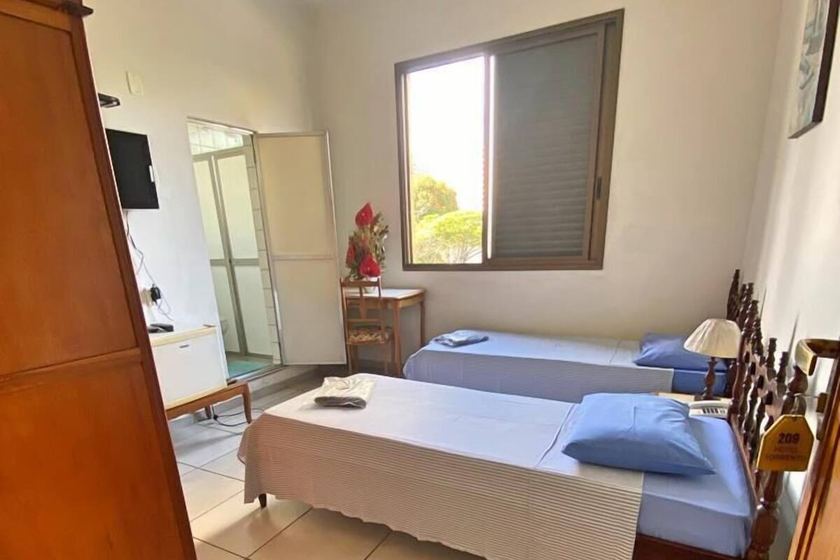Une chambre avec deux lits simples, une armoire en bois, un bureau avec une chaise et une fenêtre avec des stores.