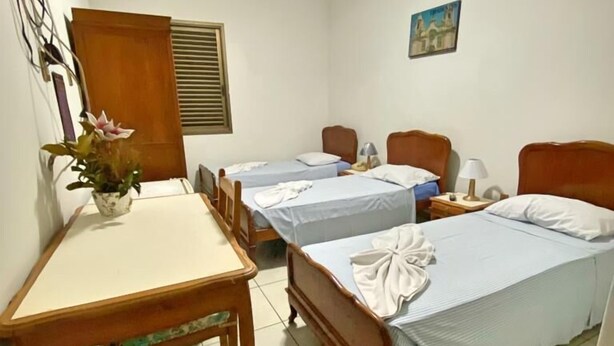 Habitación de hotel con dos camas individuales, un escritorio de madera, una silla, un ventilador de techo y una mesita con un jarrón de flores.