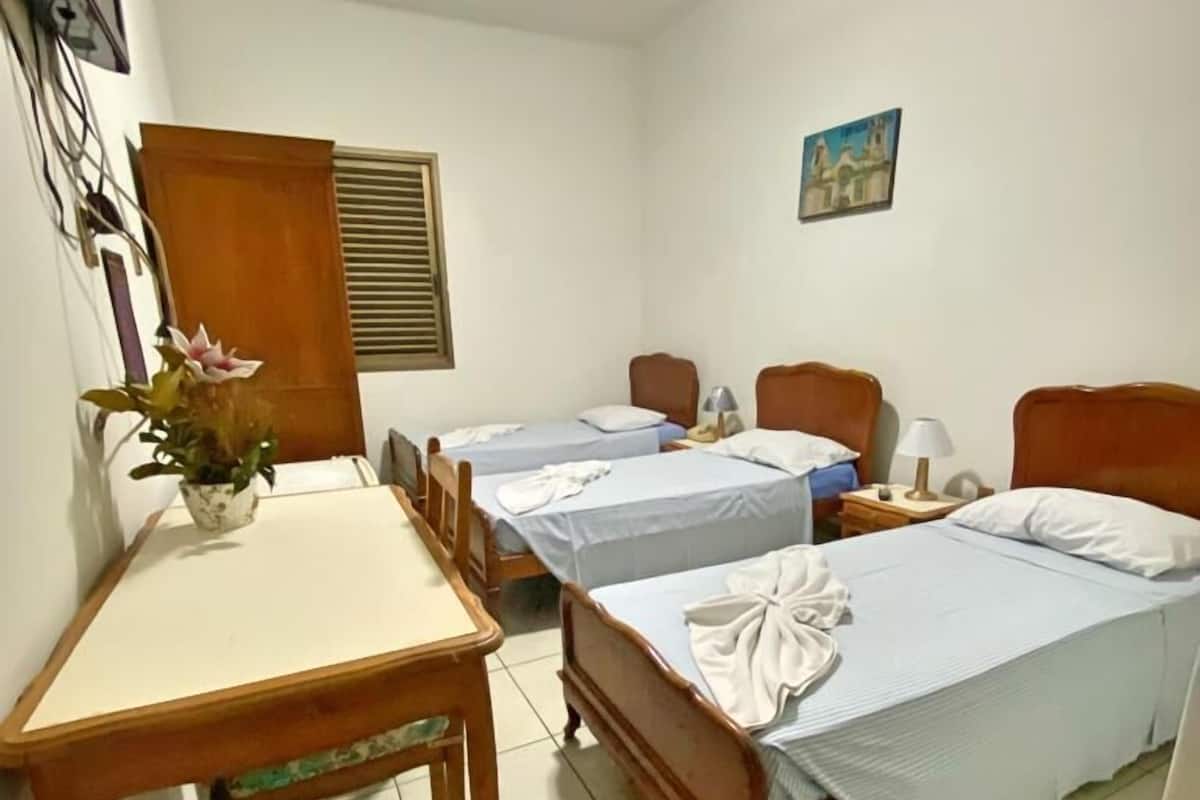 Une chambre d’hôtel avec deux lits simples, un bureau en bois, une chaise, un ventilateur de plafond et une petite table sur laquelle est posé un vase de fleurs.