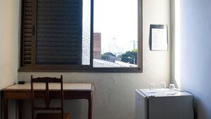 Una habitación con escritorio de madera, una silla, ventana con persianas y un refrigerador pequeño.