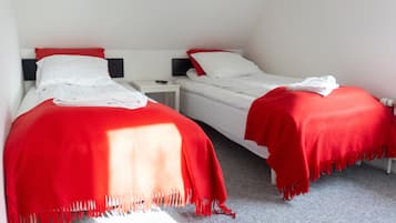 Two single beds with red blankets and white pillows, a small bedside table with a remote control, and a grey carpeted floor.