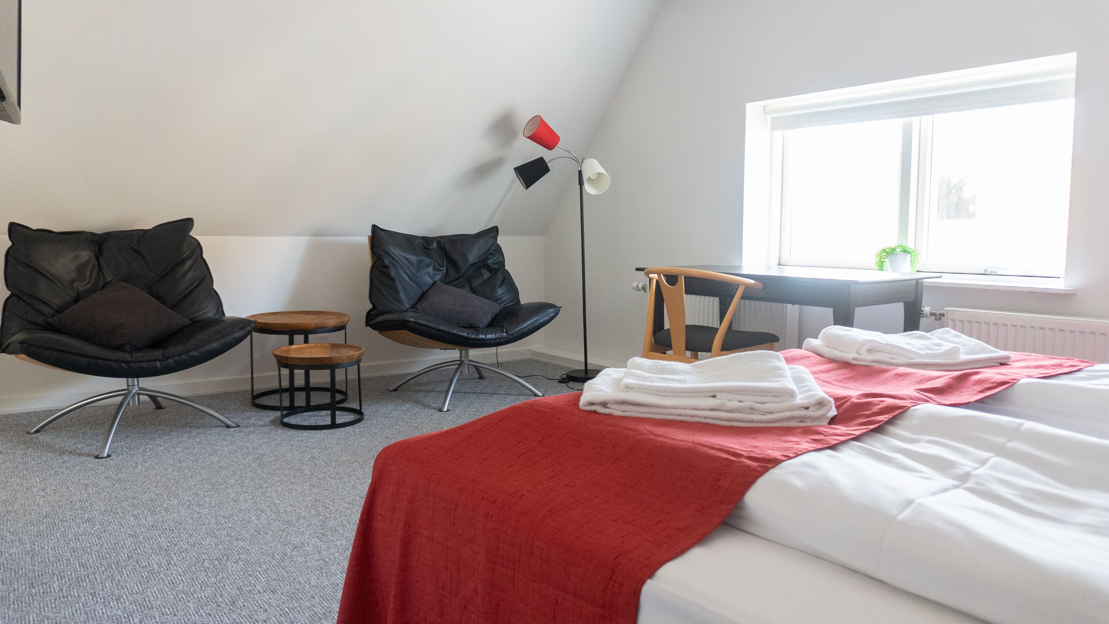 A hotel room with a bed, two chairs, a small table, a floor lamp, and a window with a view of greenery.