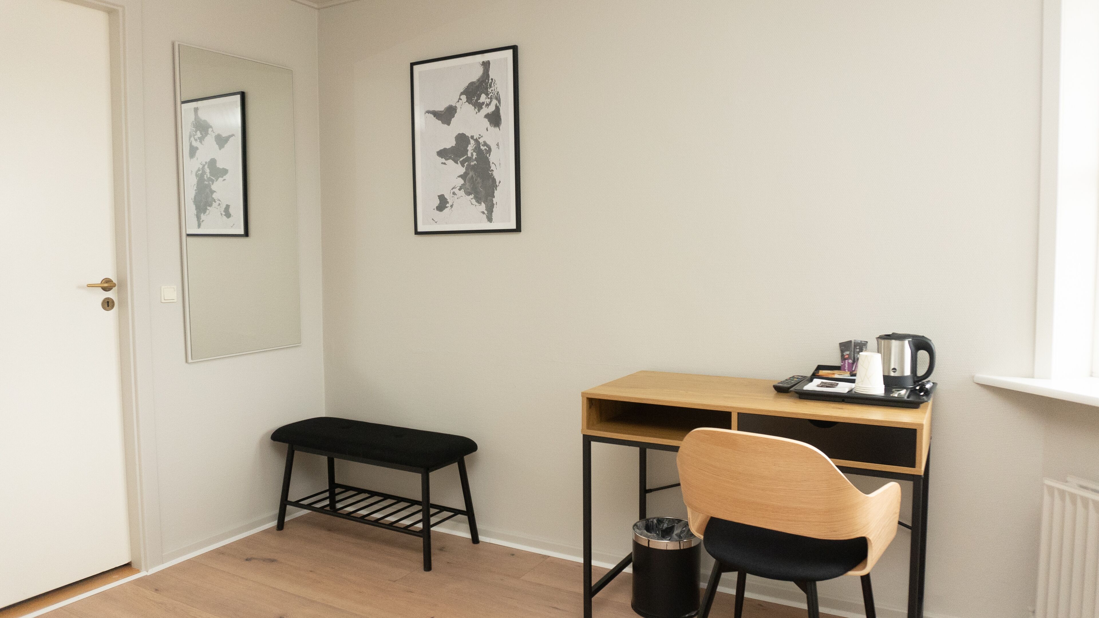 A small, tidy room with a wooden desk, a black chair, a small bench, and a trash can. There are framed pictures on the wall and a window with blinds.