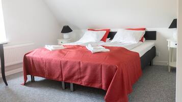 Una cama bien hecha con una colcha roja, almohadas blancas y un cabecero negro. Hay dos mesitas de noche con lámparas, un radiador blanco y una alfombra gris.