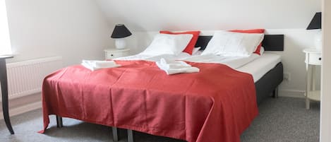 Una cama bien hecha con una colcha roja, almohadas blancas y un cabecero negro. Hay dos mesitas de noche con lámparas, un radiador blanco y una alfombra gris.