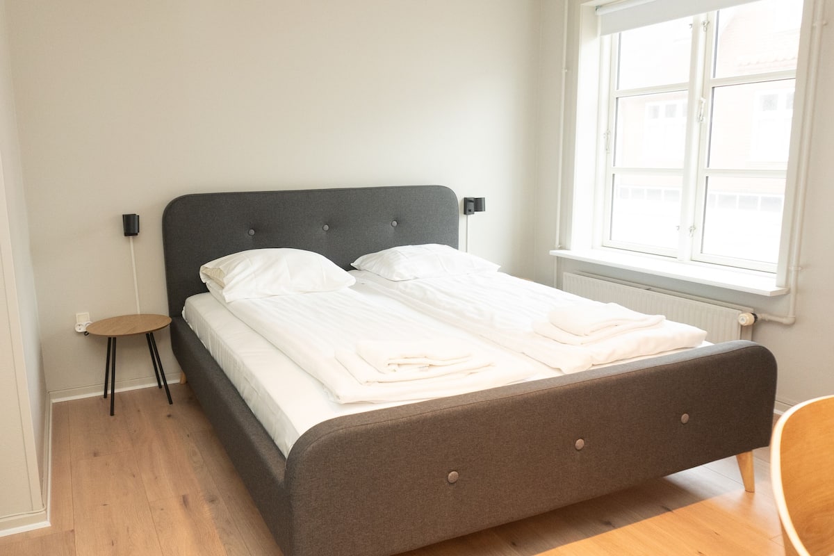 A neatly made bed with white linens, a small round table, and a chair in a room with wooden flooring and a window.