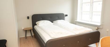 Una cama bien hecha con sábanas blancas, una mesita redonda y una silla en una habitación con suelo de madera y una ventana.