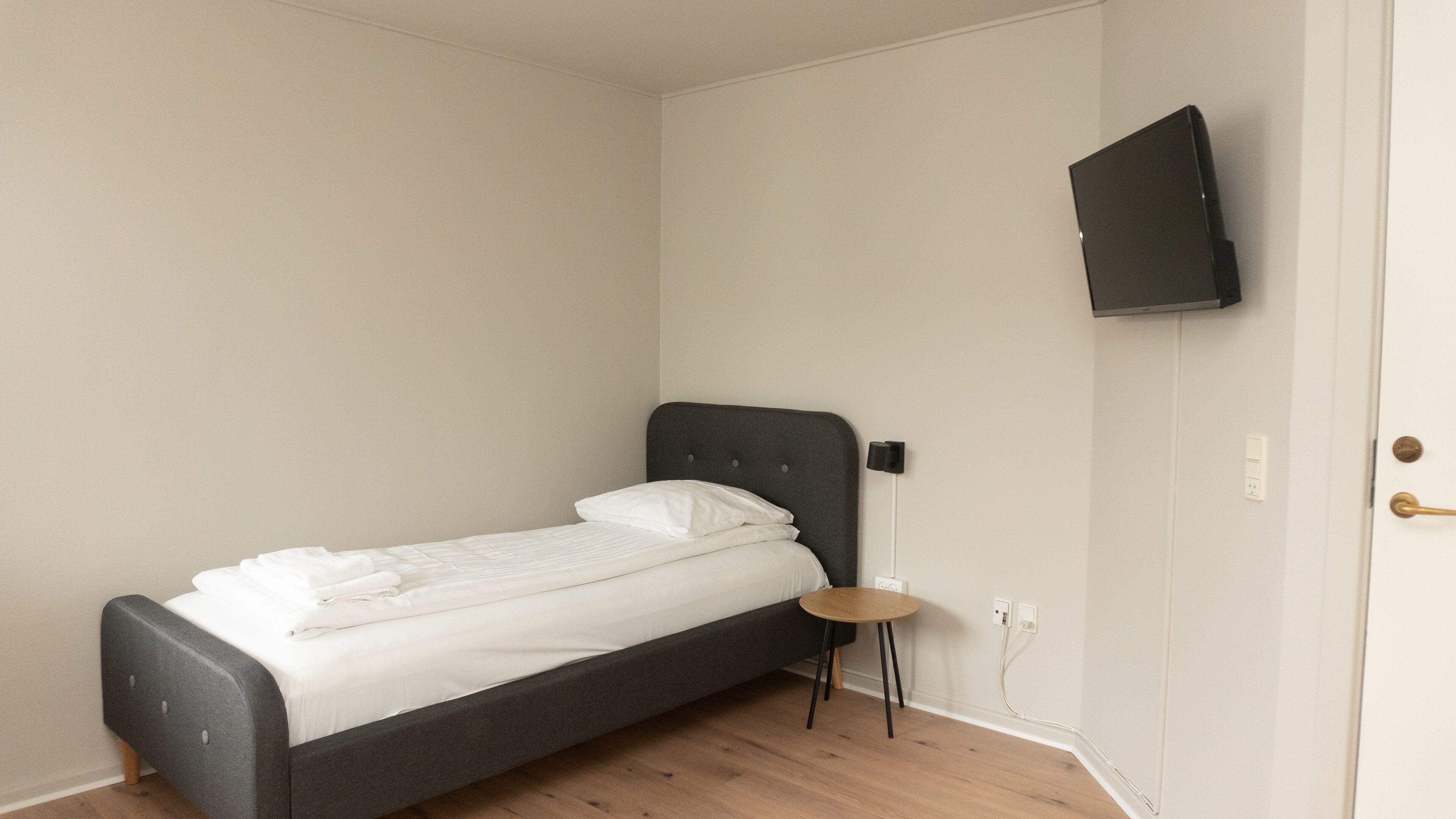 A single bed with a dark headboard, a small round table, and a TV mounted on the wall.