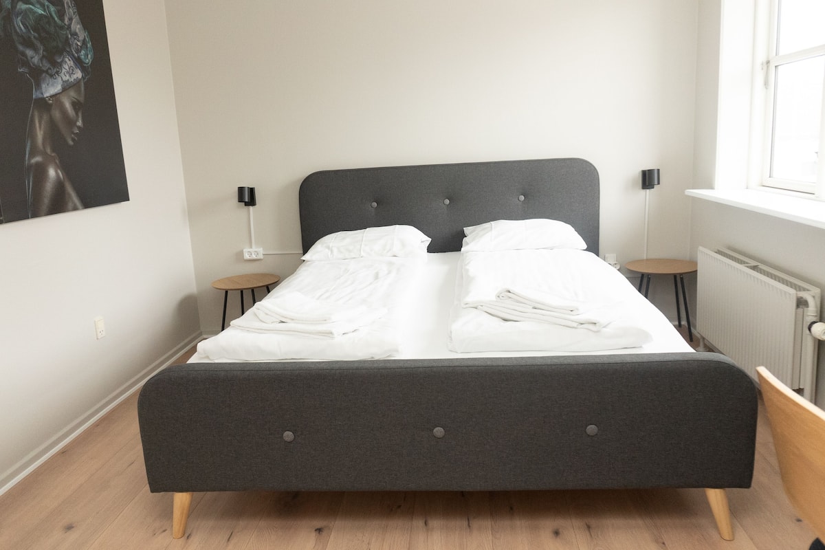A neatly made bed with white linens, a dark grey headboard, and two bedside tables with lamps.