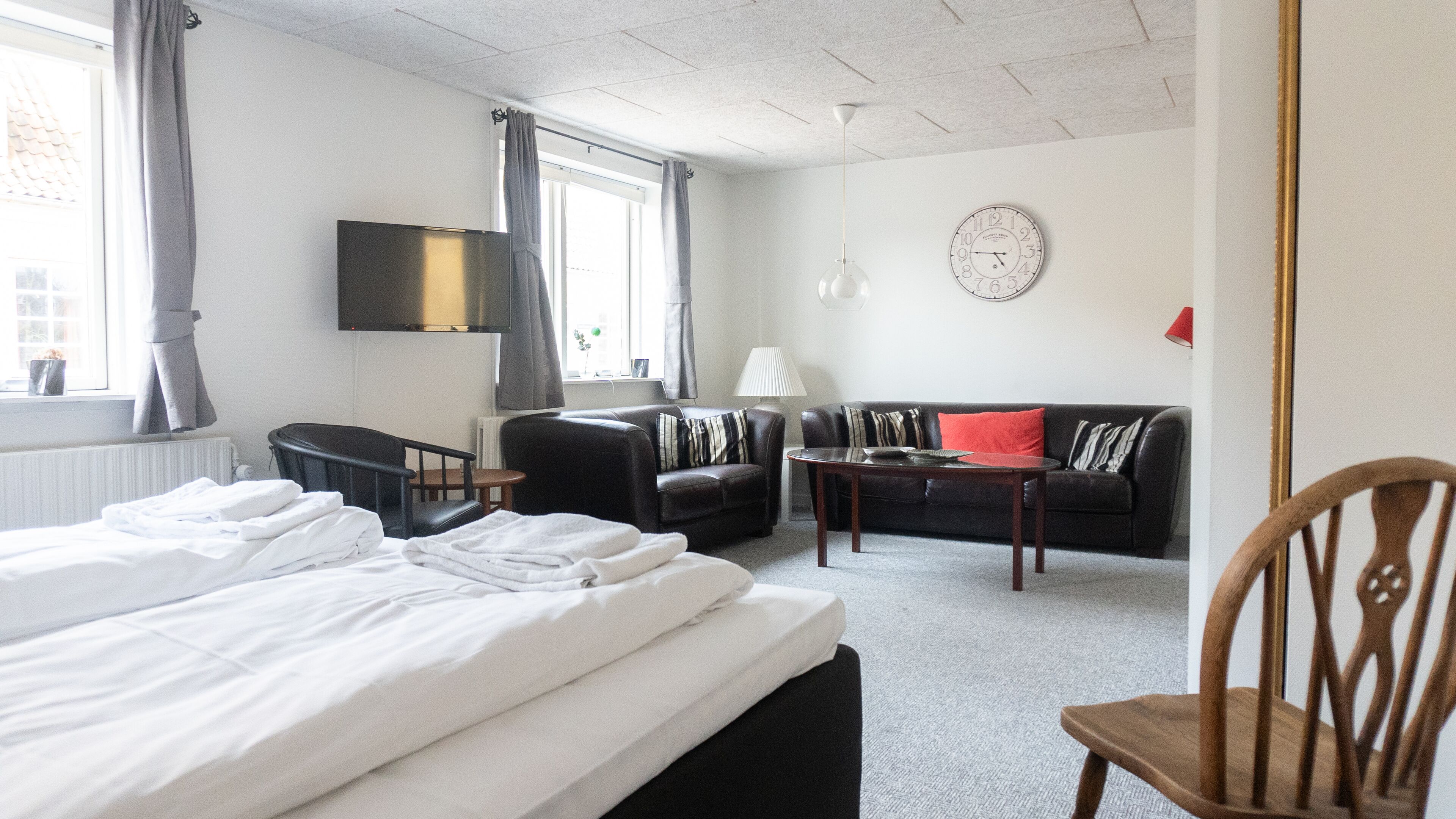 A hotel room with a bed, a sofa, a chair, a TV, a lamp, and a clock on the wall.