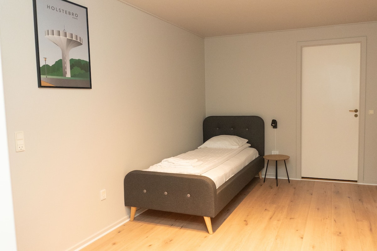 A small, single-bed room with a grey headboard, a small bedside table, and a door to the right.