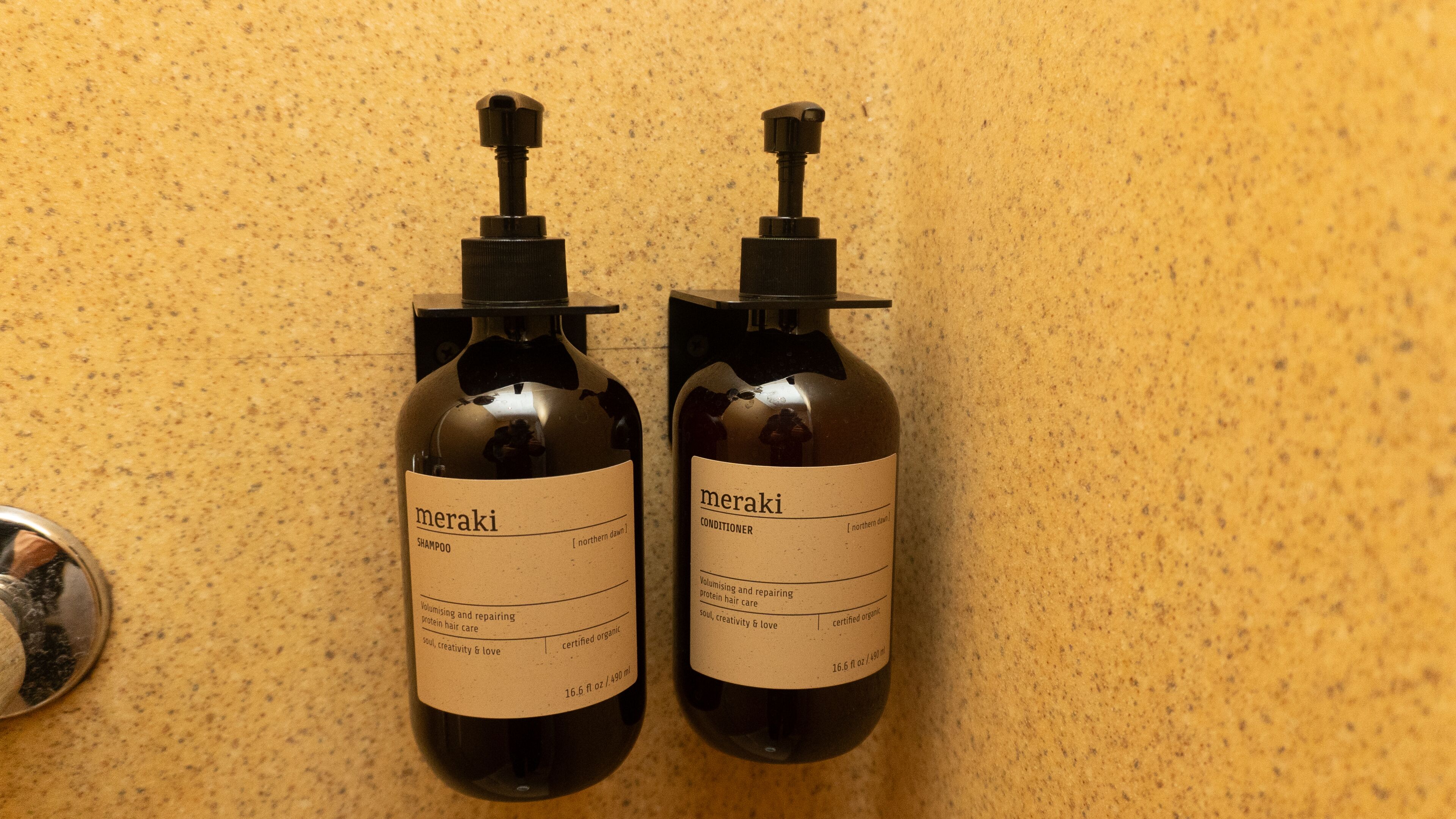 Two bottles of Meraki shampoo and conditioner on a speckled countertop.