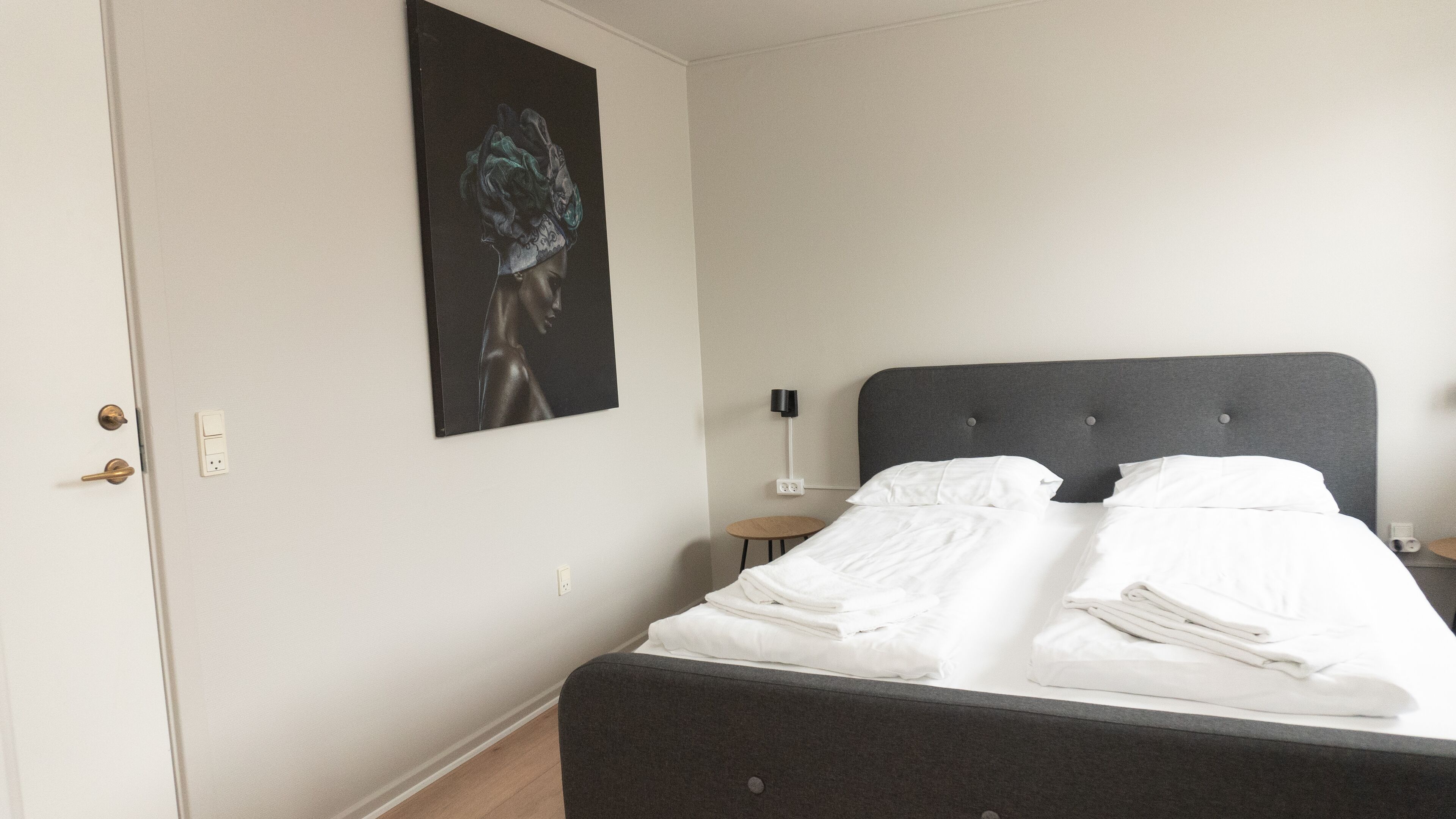 Ein ordentlich bezogenes Bett mit weißen Leinen, einem dunkelgrauen Kopfteil und einem kleinen Nachttisch mit Lampe.
