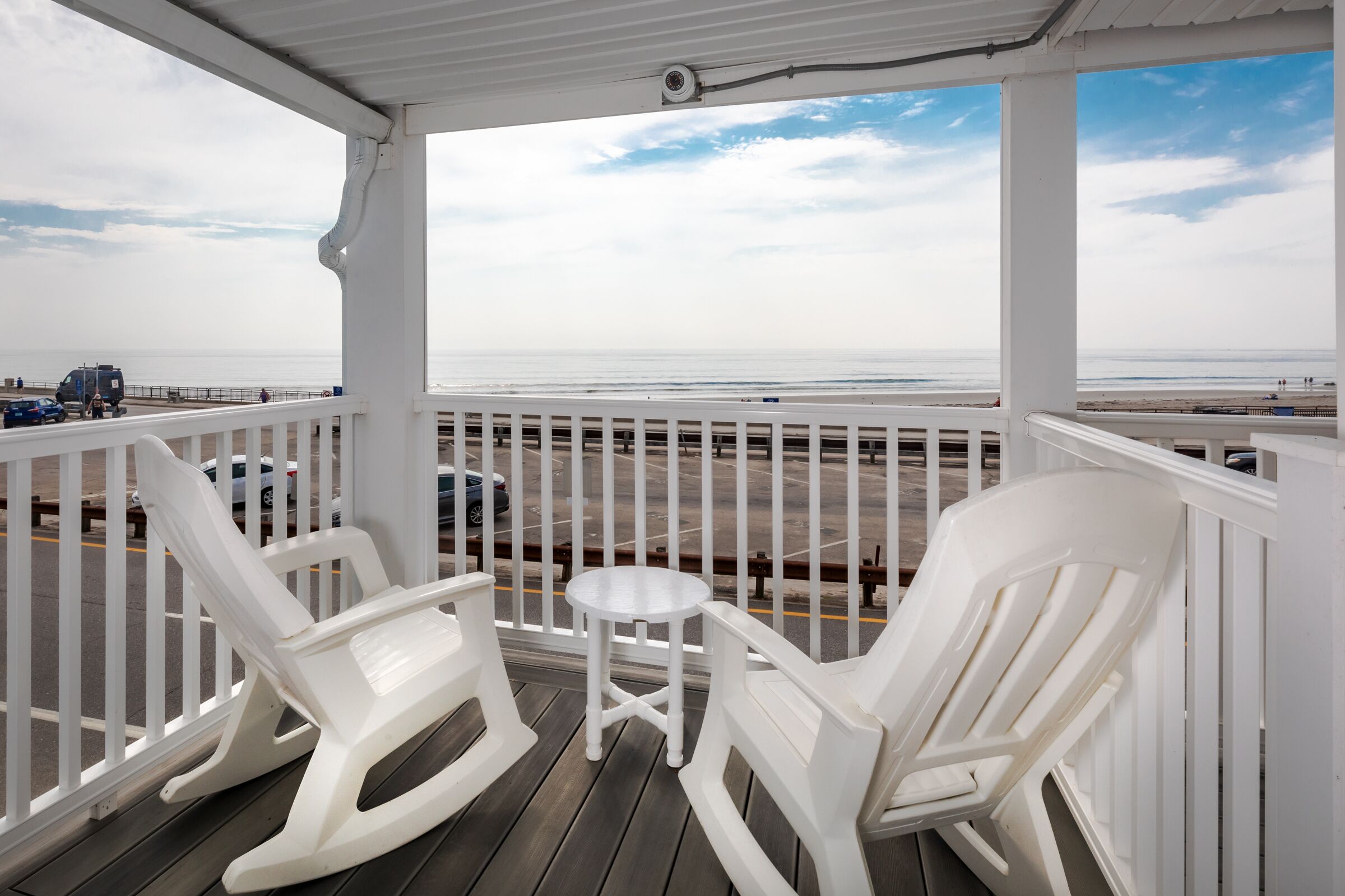 Une terrasse couverte avec deux fauteuils à bascule blancs et une petite table, d’où l’on a vue sur une plage et une route fréquentée par des véhicules.