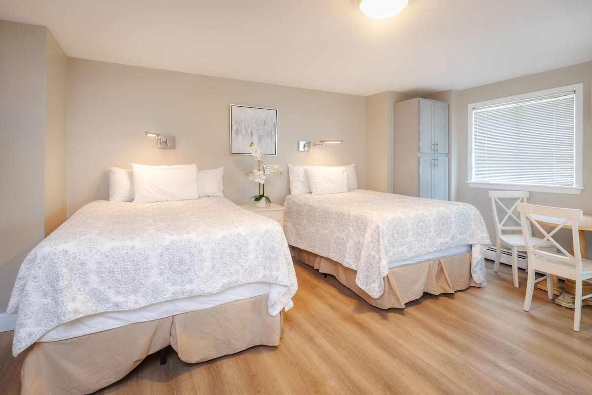 A room with two beds, a chair, a window with blinds, and a wall-mounted light fixture.
