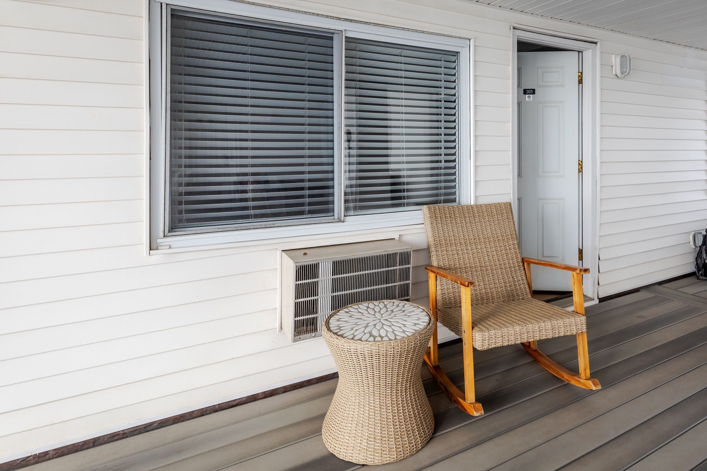A porch with a rocking chair and a woven stool, a white door, and a window with blinds.