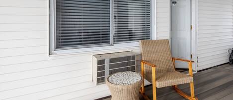 A porch with a rocking chair and a woven stool, a white door, and a window with blinds.