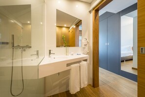 Un baño moderno con ducha de vidrio, un lavamanos blanco y un espejo encima. Hay una toalla colgada en la pared y una puerta de madera que da a un dormitorio.