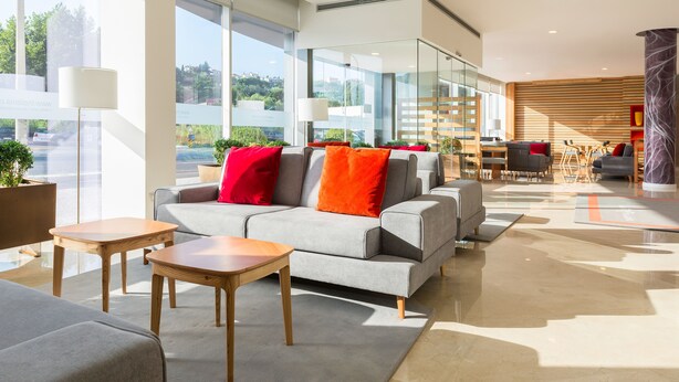 Un moderno vestíbulo de hotel con un sofá gris, mesas de madera y vistas a la ciudad a través de amplias ventanas.