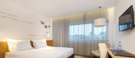 Una habitación de hotel con cama, escritorio, silla, televisión y ventana con cortinas.