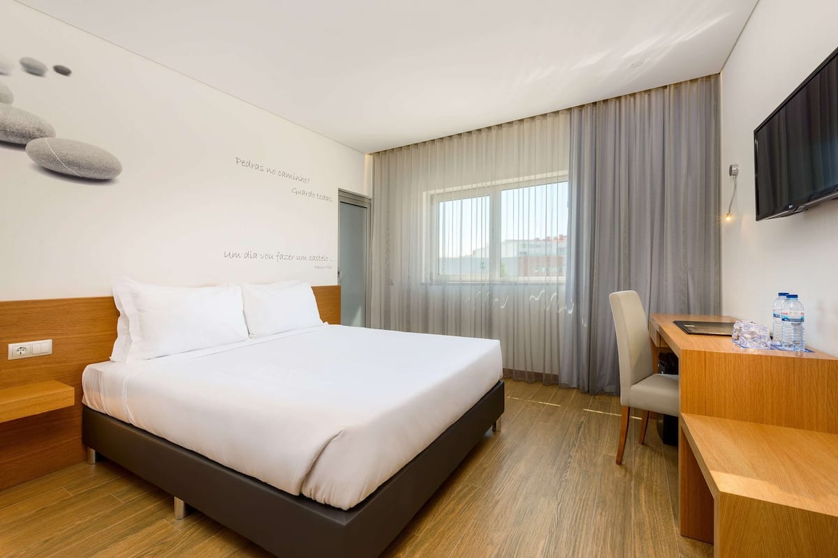Une chambre d’hôtel avec un lit, un bureau, une chaise, une télévision et une fenêtre avec des rideaux.