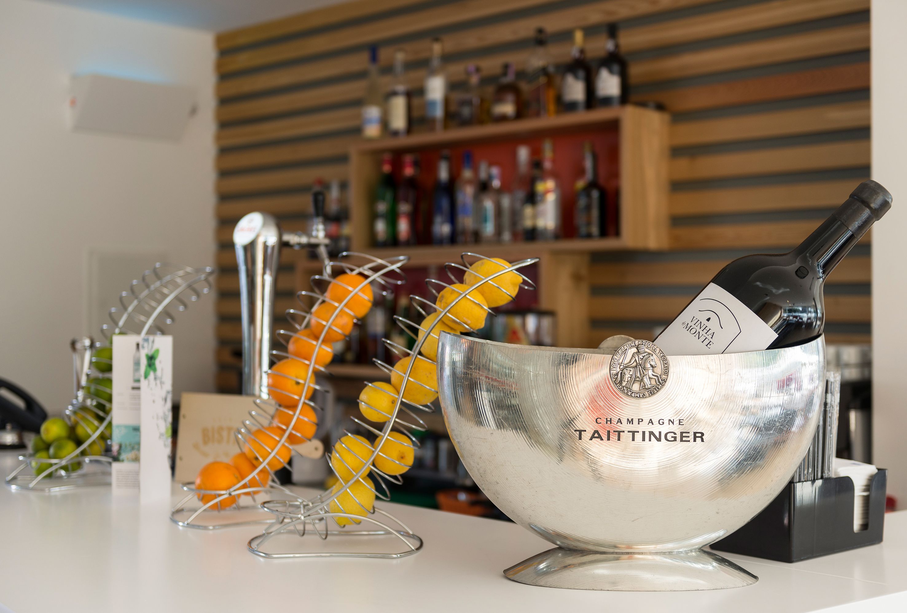 Una zona de bar con un cubo de champán que dice "Champagne Taittinger" y una botella de vino. También hay cestas de fruta y una vinoteca al fondo.