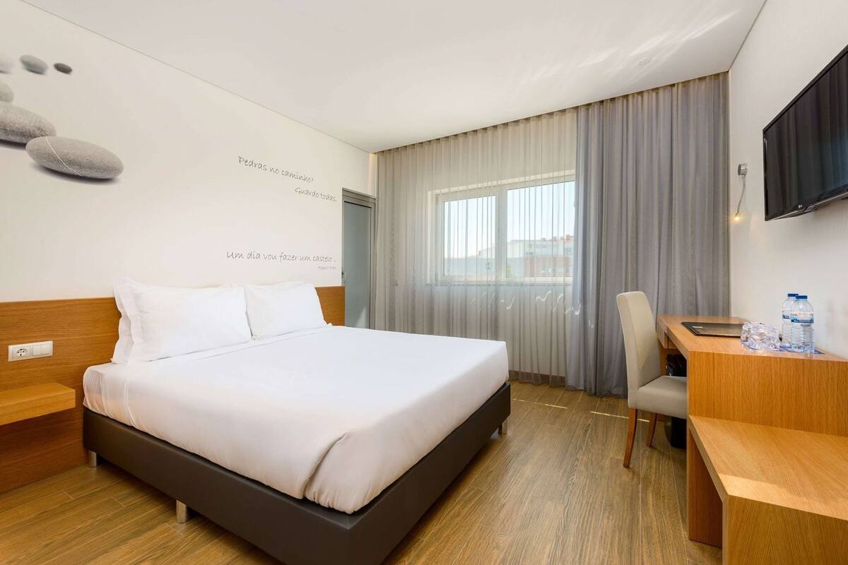 A hotel room with a bed, a desk, a chair, a TV, and a window with curtains.