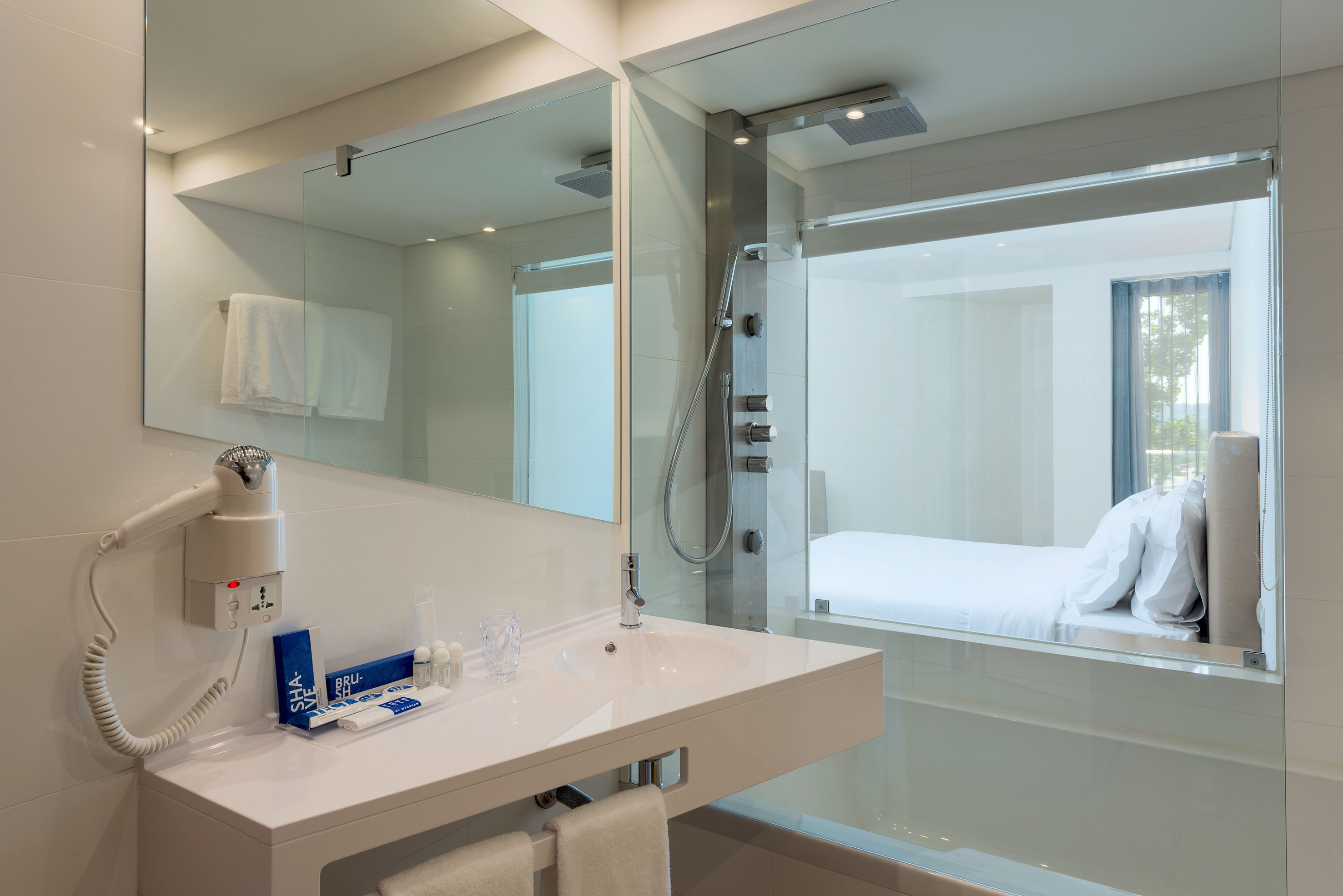 A modern bathroom with a large mirror, a white sink, a hairdryer, and a glass shower enclosure.
