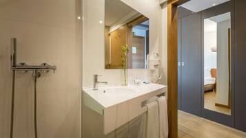 A modern bathroom with a white sink, mirror, and shower. There is a towel hanging on the wall and a wooden door leading to a bedroom.