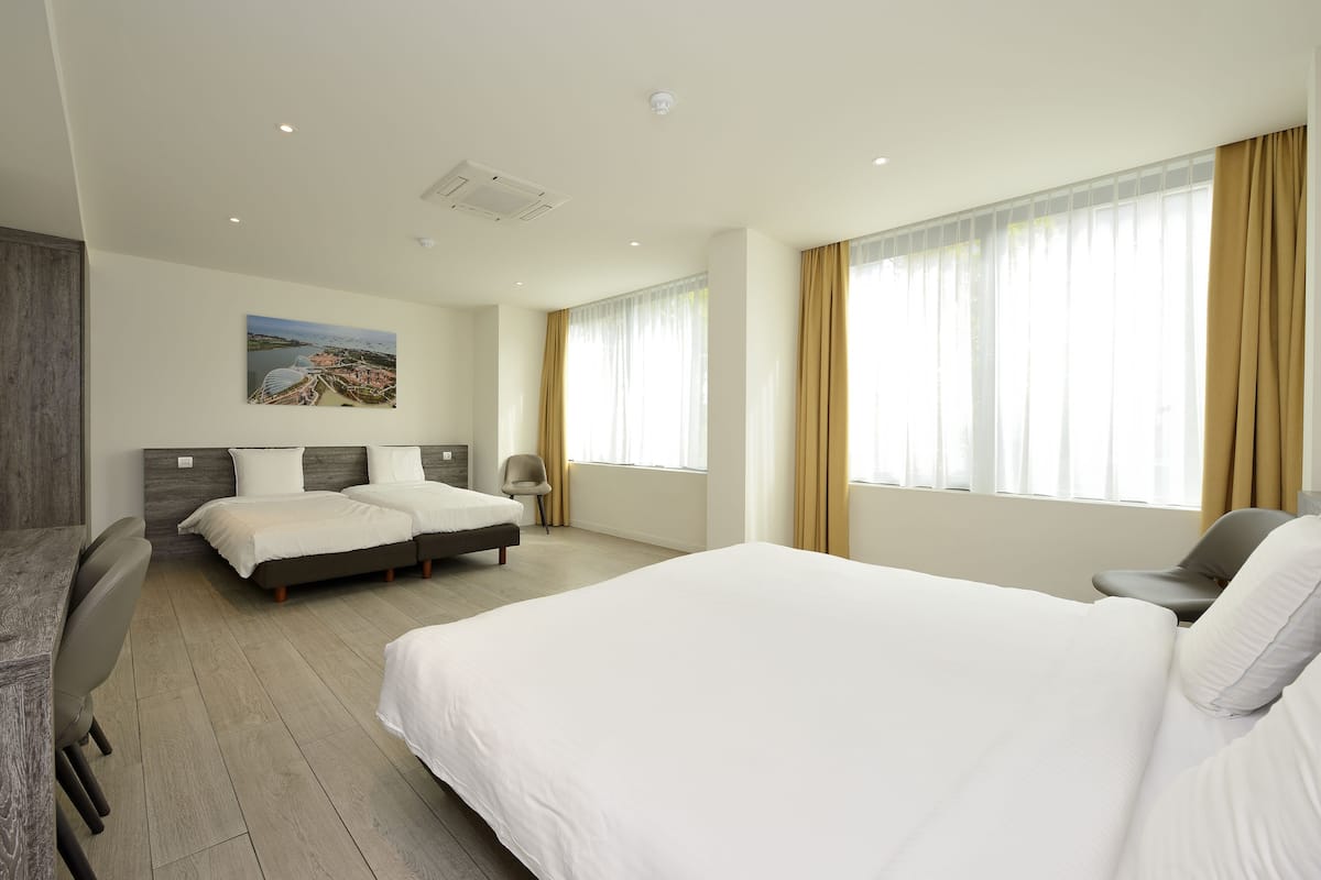 Habitación de hotel con una cama grande, dos camas más pequeñas, un escritorio y una silla. Hay un cuadro en la pared y una ventana con cortinas.