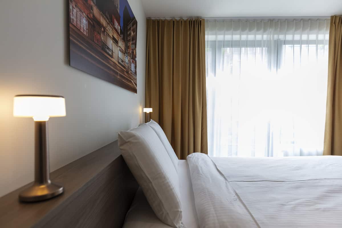 Una habitación de hotel con una cama, dos lámparas de noche, un cuadro en la pared y una ventana con cortinas.