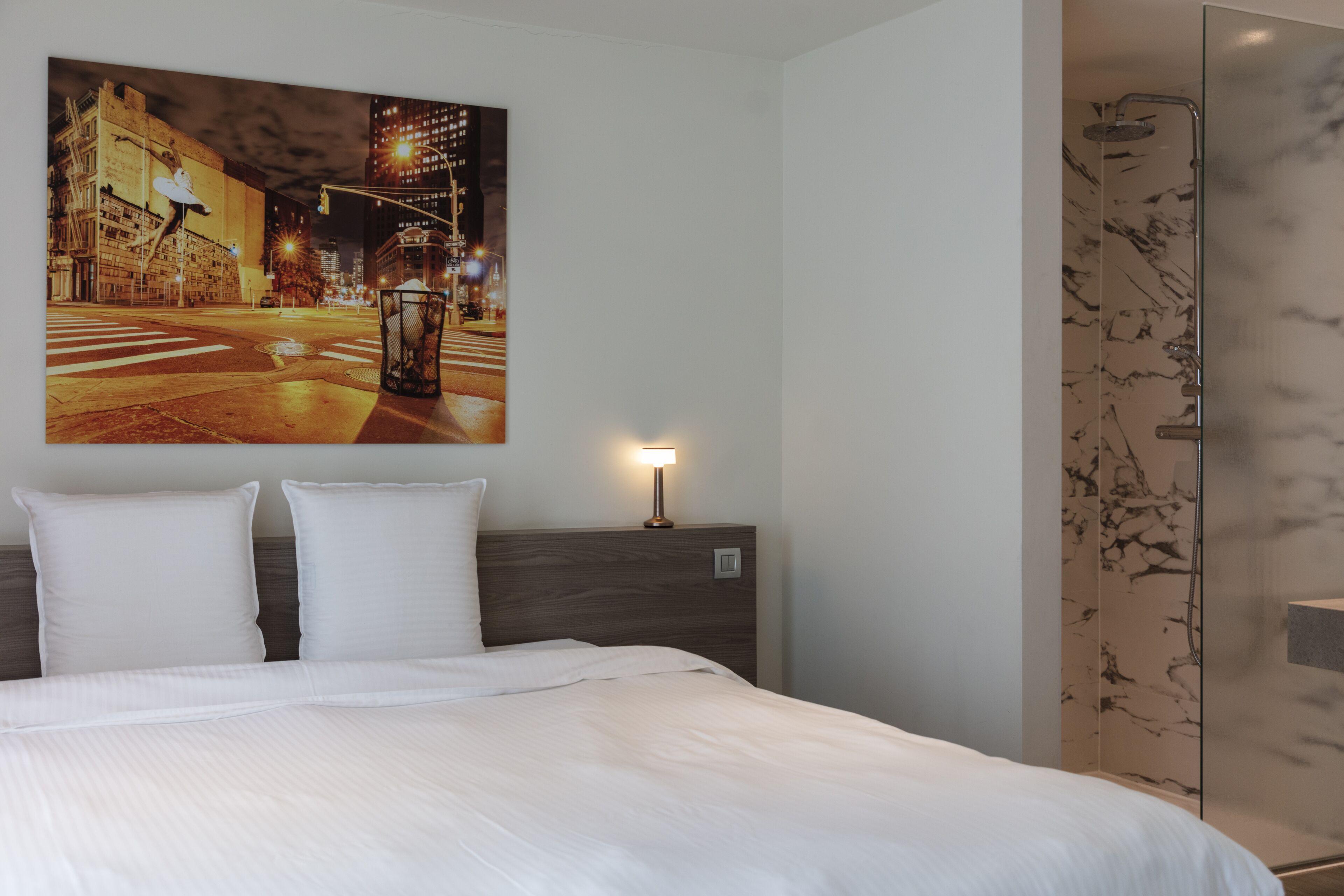 Una habitación de hotel con una cama grande, un cuadro de la ciudad nocturna, una mesita de noche con lámpara y un box de ducha de mármol.
