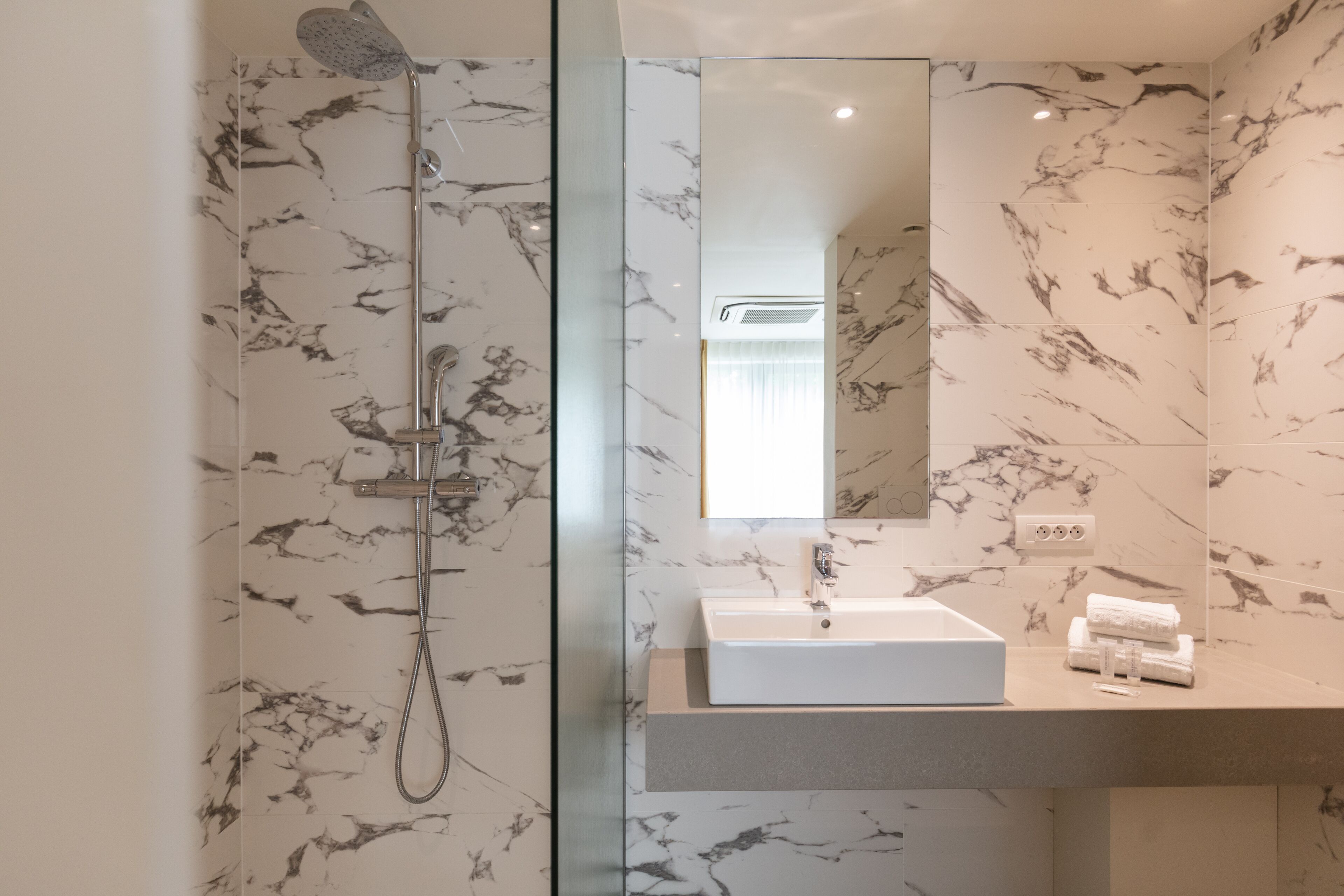 Une salle de bain moderne avec une douche à parois de verre, un pare-baignoire en marbre et un grand miroir au-dessus d’un lavabo blanc.