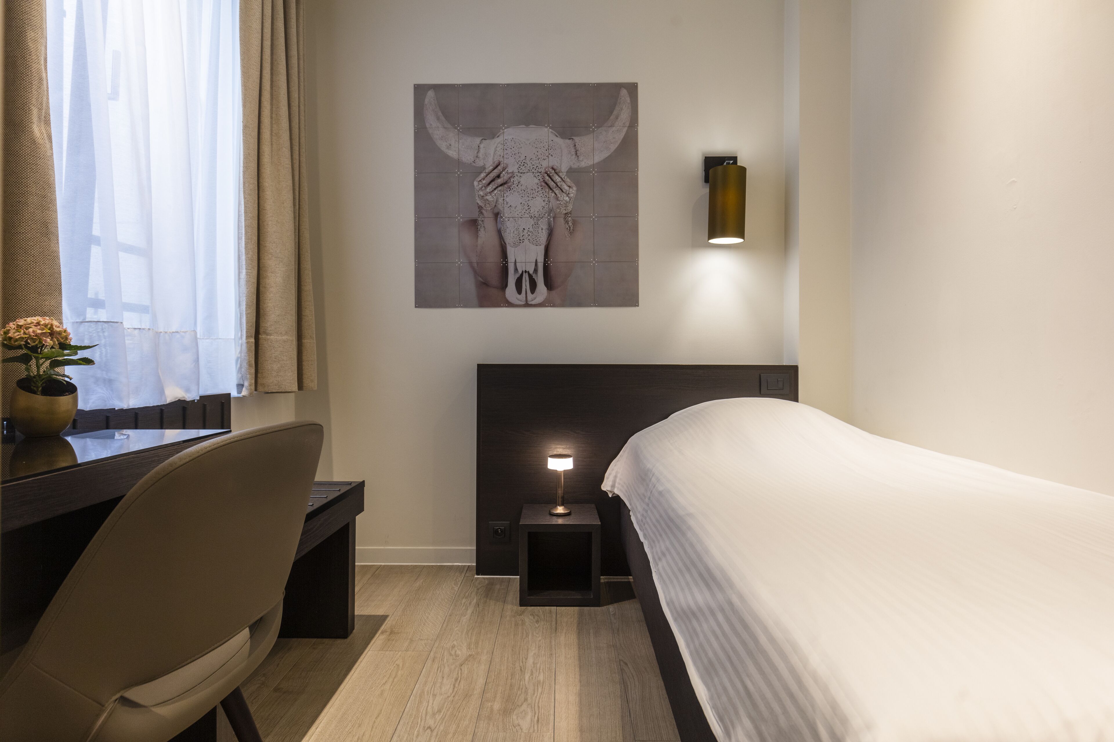 A hotel room with a bed, a desk, a chair, a nightstand with a lamp, and a wall-mounted artwork of a bull's head.