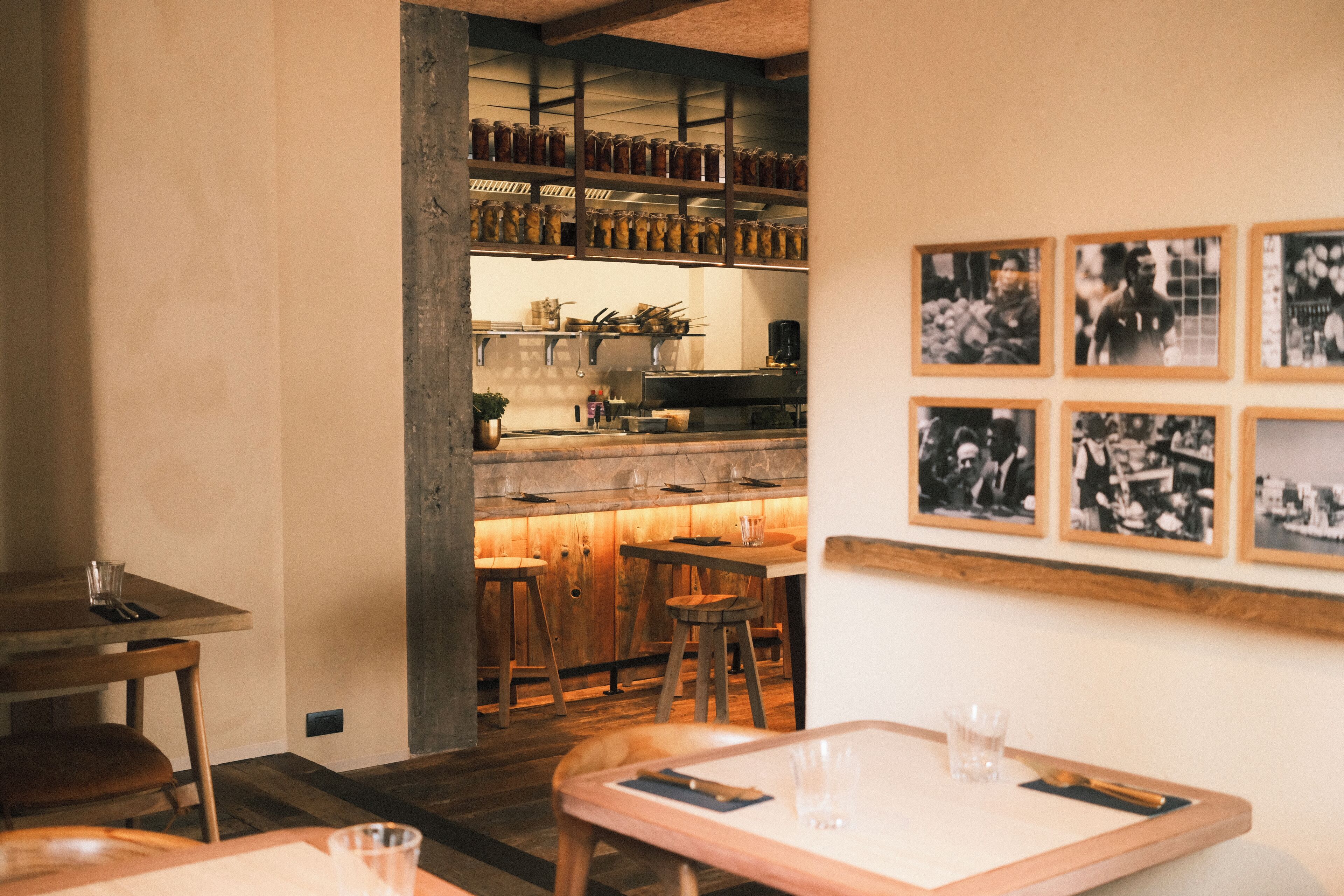A restaurant interior with wooden tables and chairs, a bar area with shelves of jars, and framed black-and-white photographs on the wall.
