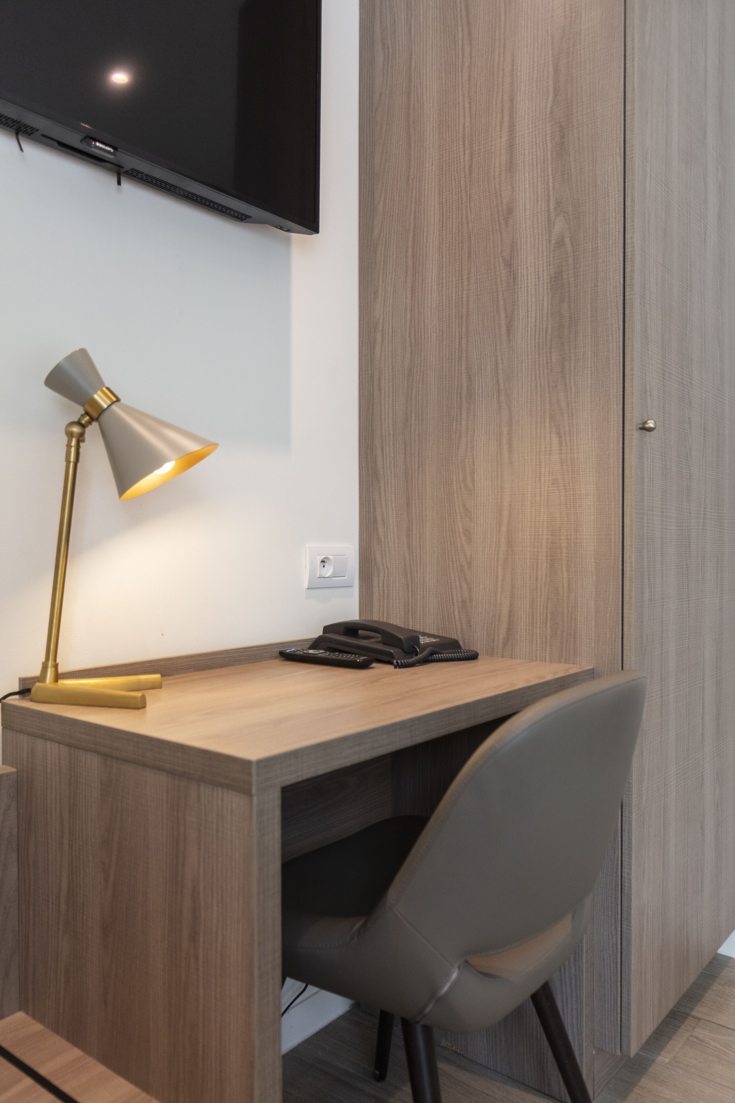 A modern office space with a wooden desk, a grey office chair, a desk lamp, a black telephone, and a mounted flat-screen TV.