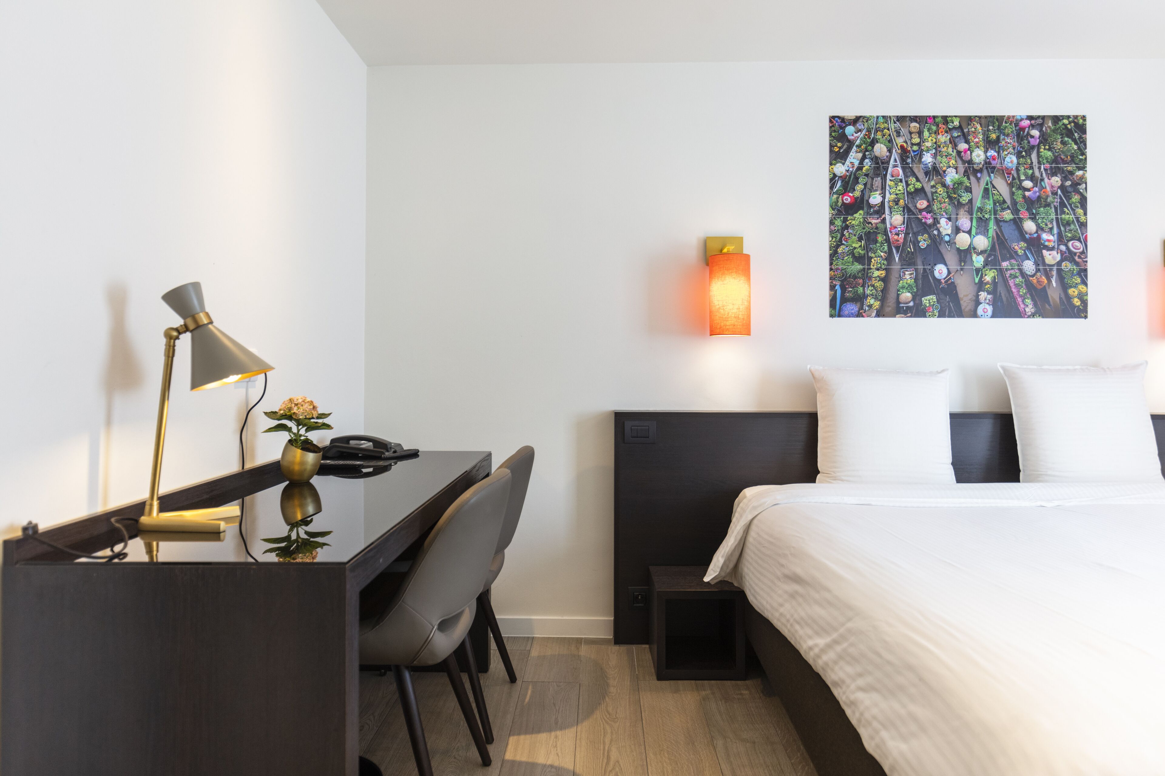 A modern hotel room with a desk, chair, bed, and a wall-mounted artwork.