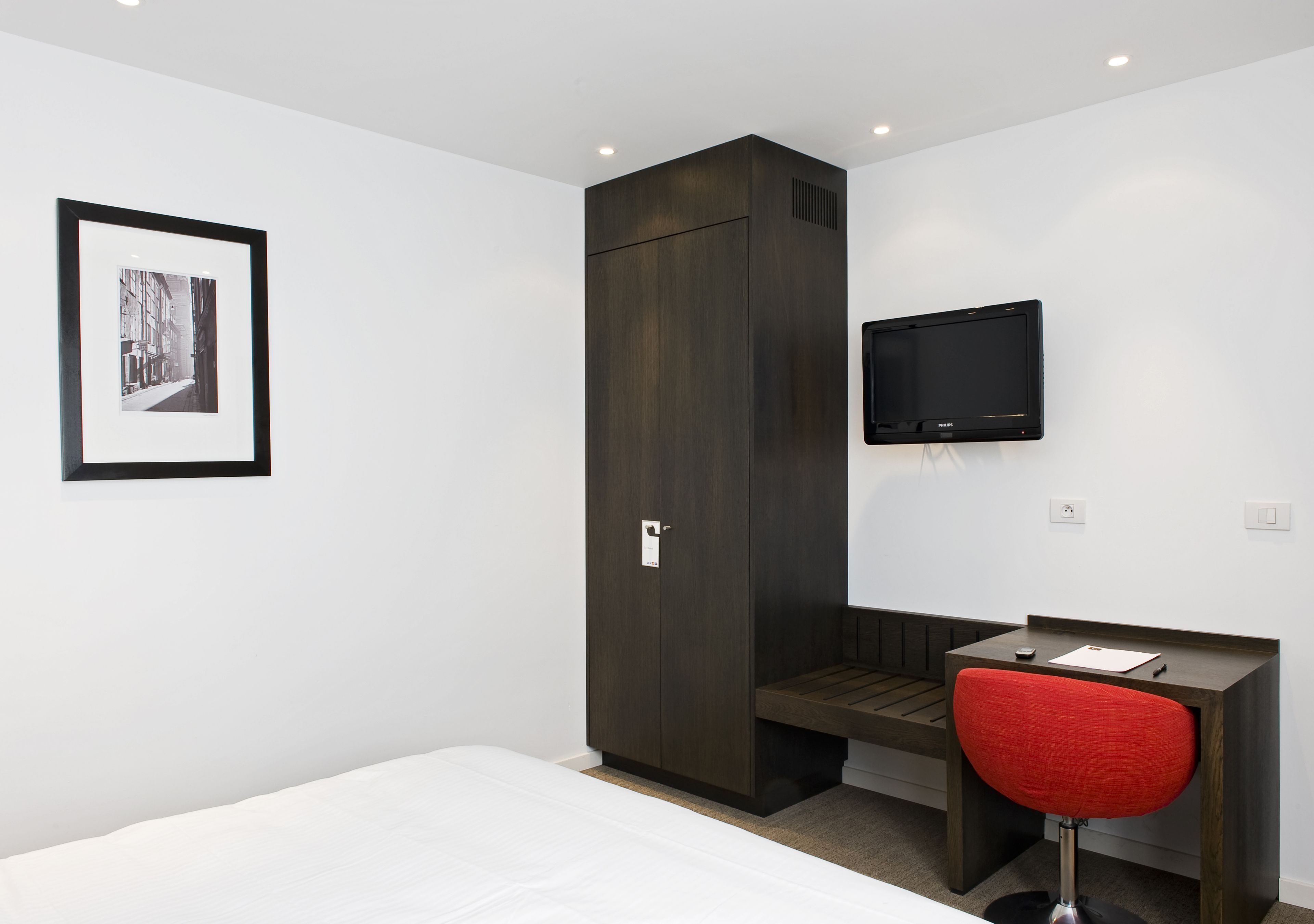 A hotel room with a bed, a desk with a chair, a wardrobe, a flat-screen TV, and a framed picture on the wall.