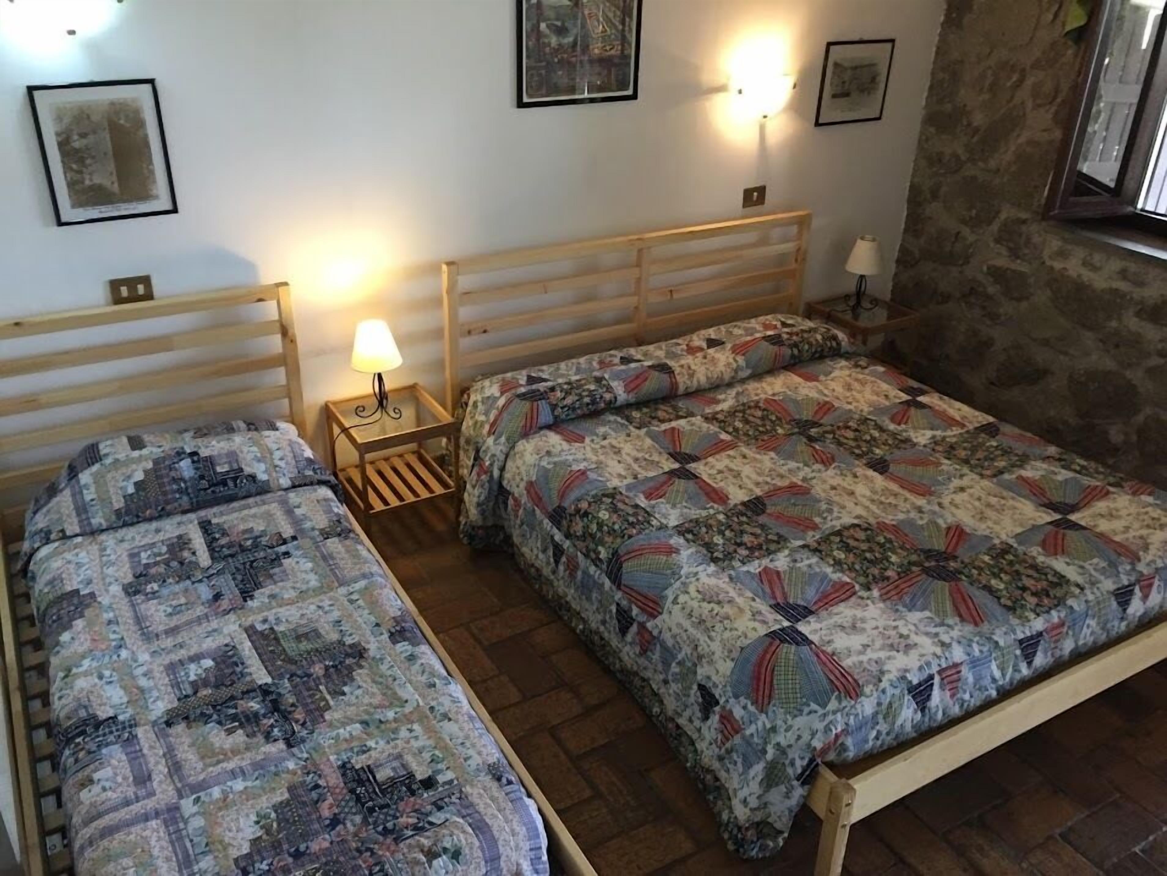 A room with two wooden beds, each with a quilted bedspread. There are wall-mounted lights and framed pictures on the walls.