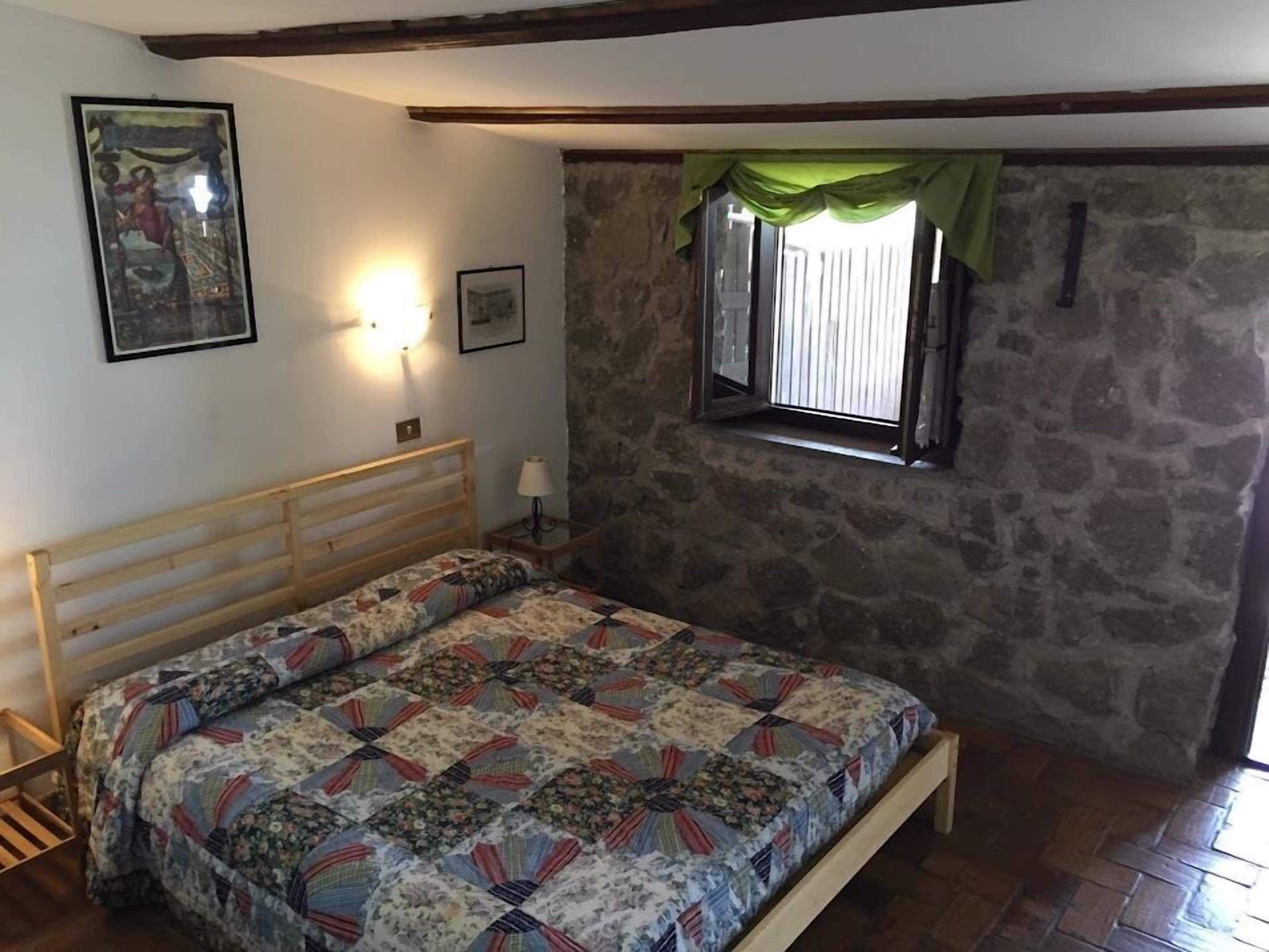 Una camera da letto con un letto di legno, un muro in pietra, una finestra con tenda verde e due quadri appesi al muro.