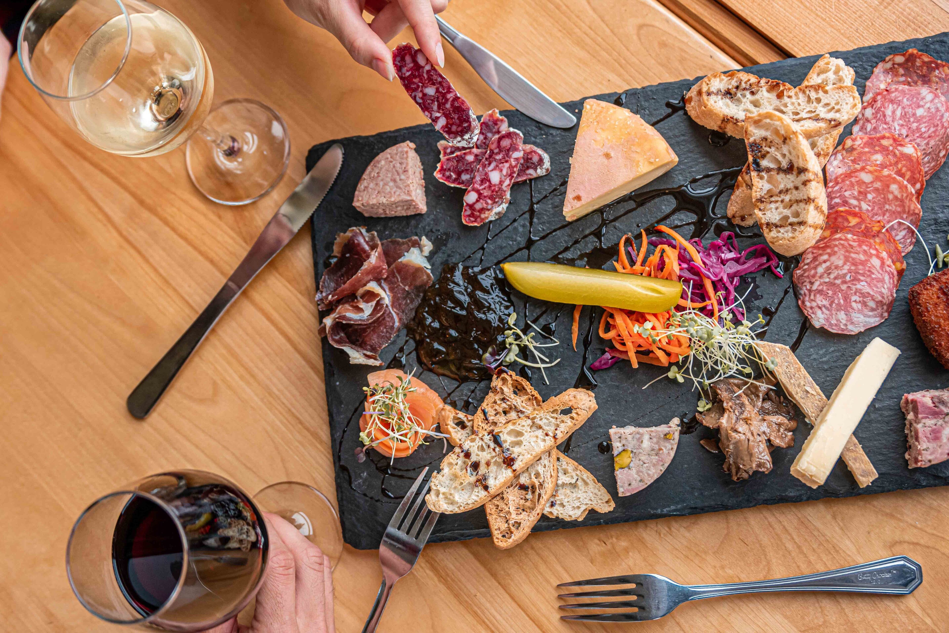 A charcuterie board with various meats, cheeses, and pickles on a slate board, accompanied by wine glasses and cutlery.