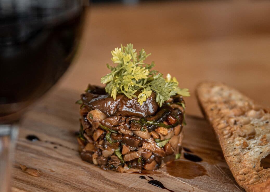 Une terrine de champignons et d’herbes aromatiques, servie sur une planche en bois, accompagnée d’une tranche de pain.