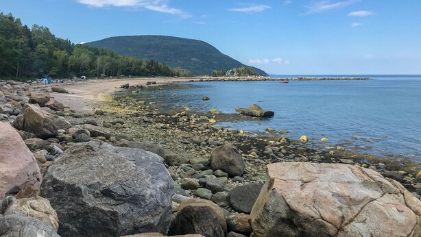 Una playa rocosa con tiendas de campaña y un bote, rodeada de árboles y una colina.