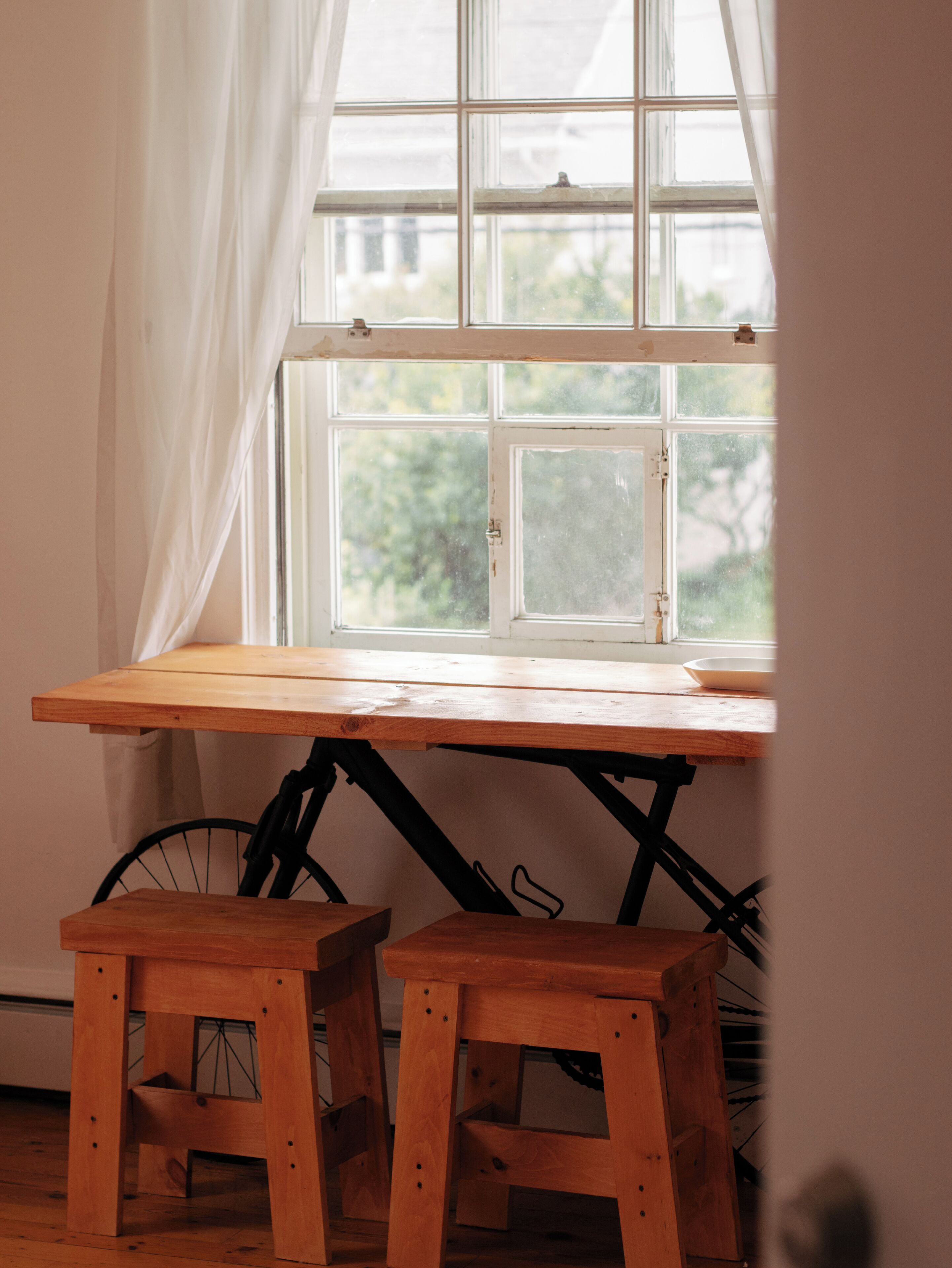 A wooden table with a window above it, two wooden stools, and a bicycle in the background.