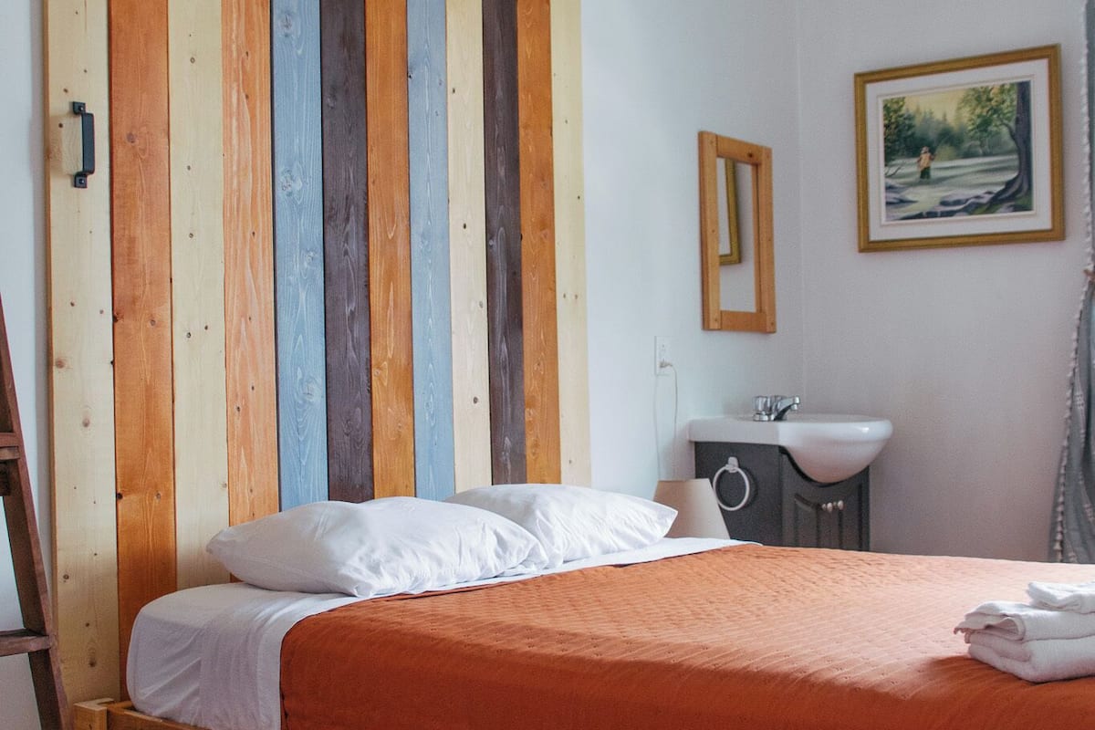 Una cama individual con estructura de madera y colcha naranja, un armario de madera con puerta corredera, un lavabo blanco y un cuadro en la pared.